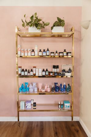 Gold metal shelf against a blush-pink wall displaying assorted skincare and haircare bottles, jars and pastel packaging with two potted trailing plants on top — chic retail beauty display.