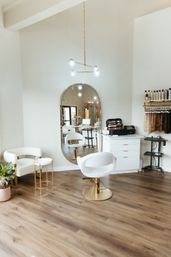 Bright modern hair salon interior with white-and-gold styling chair, large oval mirror, wood flooring, product shelves and hanging hair extensions in a chic boutique beauty studio