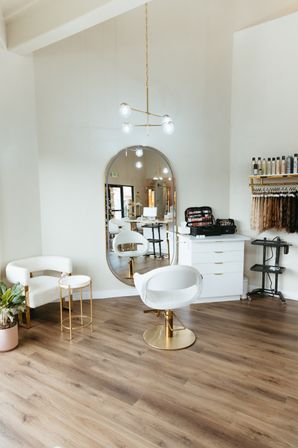 Bright modern hair salon interior with white-and-gold styling chair, large oval mirror, wood flooring, product shelves and hanging hair extensions in a chic boutique beauty studio