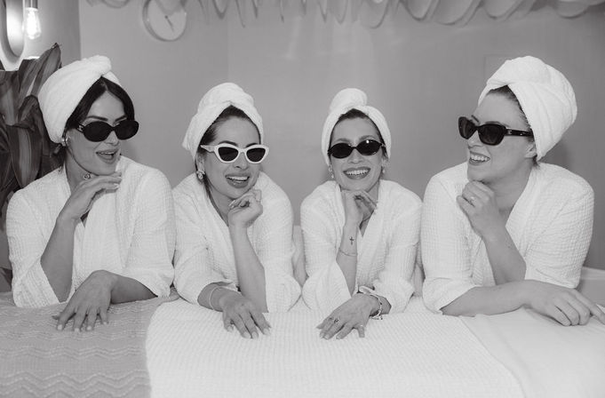 Black-and-white photo of four women in white robes and towel turbans wearing sunglasses, smiling and relaxing together on a spa bed in an indoor spa setting.