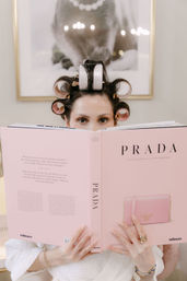 Woman with pink and white hair rollers peeking over a large pink fashion book, wearing a white robe and gold rings in a chic beauty setting.