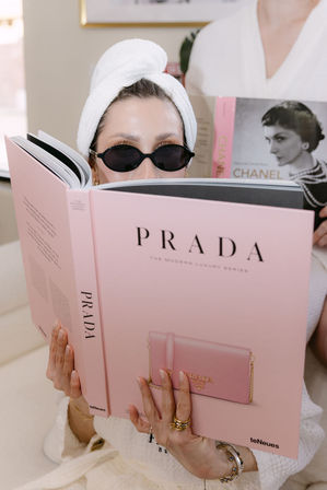 Woman in a spa robe with towel turban and black oval sunglasses reading a large pink fashion coffee-table book featuring a small pink purse on the cover; gold rings and bracelets on her hands and another classic fashion book peeking behind.
