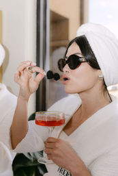 Woman in white spa robe and towel turban wearing round black sunglasses, holding a pink cocktail coupe and lifting two blackberries on a toothpick to her lips — luxe spa pampering moment.