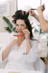 Playful woman in a white robe sitting in a salon chair with hair rollers while a stylist wraps a section, winking and sipping a pink cocktail from a coupe glass.
