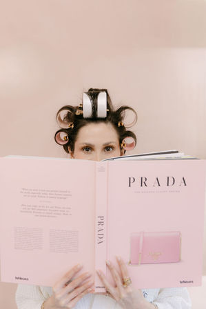 Person with pink hair rollers peeking over a large pink fashion book, manicured hands with rings holding it against a blush background — playful at-home beauty reading scene.