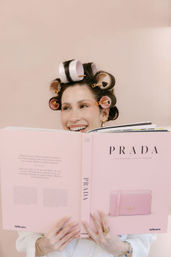 Smiling woman with pink hair rollers peeking over a large pink fashion coffee-table book showing a purse, set against a soft blush background.
