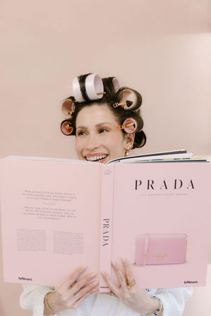 Smiling woman with pink hair rollers peeking over a large pink fashion coffee-table book showing a purse, set against a soft blush background.
