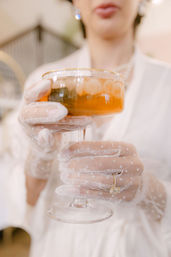 Close-up of hands in white polka-dot mesh gloves holding a gold-rimmed coupe glass of amber cocktail with ice, showcasing a delicate gold ring in an elegant, vintage-inspired setting.