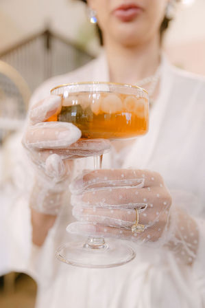 Close-up of hands in white polka-dot mesh gloves holding a gold-rimmed coupe glass of amber cocktail with ice, showcasing a delicate gold ring in an elegant, vintage-inspired setting.