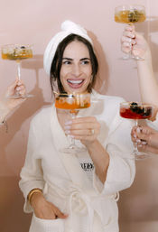 Smiling woman in a white waffle robe and towel turban cheers with a champagne coupe as friends raise colorful cocktails during a spa-day celebration.