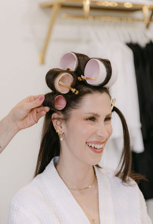 Smiling woman in a white robe getting pink hair rollers clipped into dark hair by a stylist in a bright beauty salon — close-up hair styling session with gold jewelry.