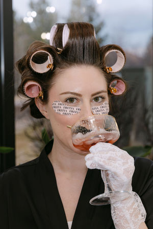 Woman with pink hair rollers and printed under-eye patches, wearing a lace glove and sipping a cocktail from a vintage coupe glass during an at-home spa beauty routine.