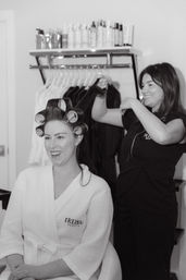 Smiling client in a white robe with large hair rollers while a stylist lifts a section of hair in a bright salon with product shelves and hanging garments in the background.