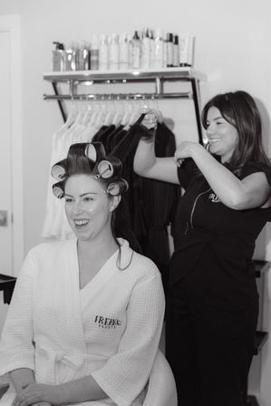 Smiling client in a white robe with large hair rollers while a stylist lifts a section of hair in a bright salon with product shelves and hanging garments in the background.