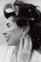 Black-and-white close-up of a woman in retro beauty prep: large hair rollers and clips, smiling with a paper eye patch, wearing a polka-dot mesh glove, pearl necklace and sparkly earring.