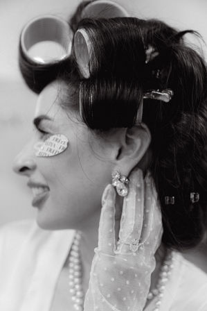 Black-and-white close-up of a woman in retro beauty prep: large hair rollers and clips, smiling with a paper eye patch, wearing a polka-dot mesh glove, pearl necklace and sparkly earring.