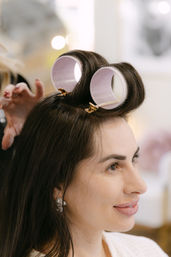 Smiling woman in a hair salon with pink velcro rollers clipped at her crown, dark hair and sparkly earrings during professional styling.
