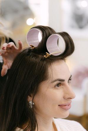 Smiling woman in a hair salon with pink velcro rollers clipped at her crown, dark hair and sparkly earrings during professional styling.