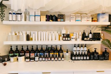 Bright spa or salon display of neatly arranged skincare products on white shelves — bottles, jars, pumps and dropper serums in organized rows.