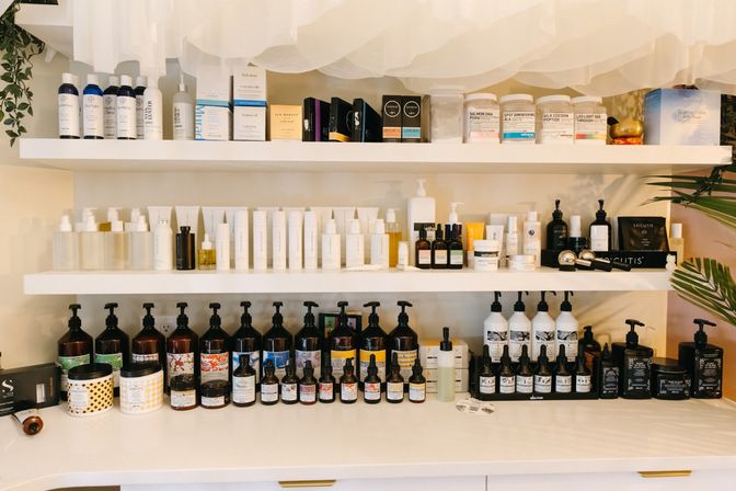 Bright spa or salon display of neatly arranged skincare products on white shelves — bottles, jars, pumps and dropper serums in organized rows.