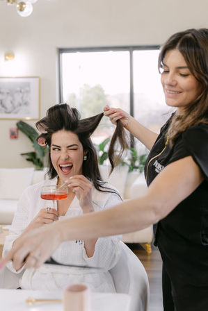 Client in a white robe winks and sips a pink cocktail while a hairstylist sets rollers and lifts her hair in a bright modern salon.