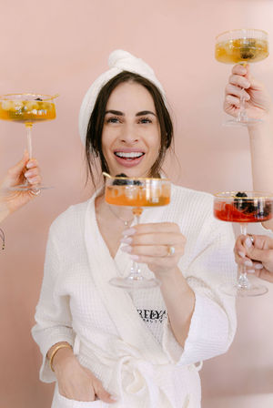 Smiling woman in a white spa robe and towel turban toasting with colorful coupe cocktails against a pale pink backdrop