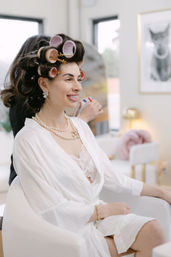 Smiling woman in a white robe and lace camisole sitting in a modern salon chair with pink hair rollers, pearl necklace and earrings as a stylist sprays hairspray during bridal getting-ready beauty prep