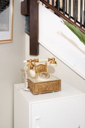Ornate gold-and-ivory vintage rotary telephone on a white cabinet in a home entryway beside a staircase with dark wood handrail and a green plant.