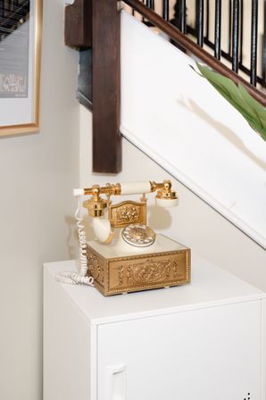 Ornate gold-and-ivory vintage rotary telephone on a white cabinet in a home entryway beside a staircase with dark wood handrail and a green plant.