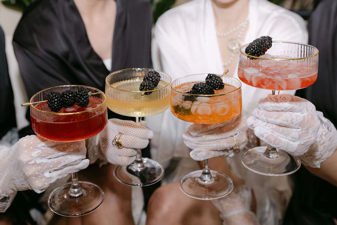 Four colorful blackberry‑garnished cocktails in coupe glasses held by hands wearing white lace gloves at an elegant indoor celebration