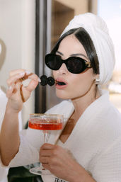 Person in a white towel turban and robe wearing black sunglasses, holding a coupe of pink cocktail and eating skewered blackberries — chic spa/resort vibe