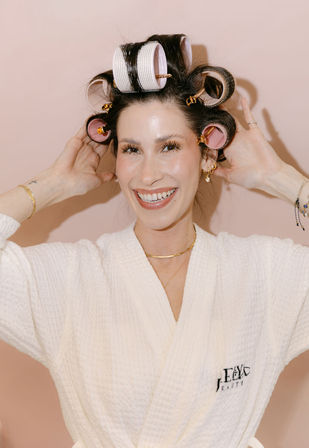 Smiling woman in a white spa robe wearing pink and white hair rollers, hands adjusting curls against a pale pink backdrop — playful beauty routine shot