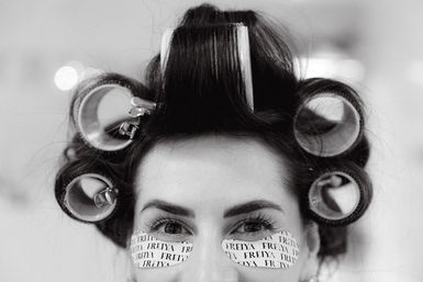 Close-up black-and-white photo of a woman wearing hair rollers and printed under-eye gel patches in a beauty salon.
