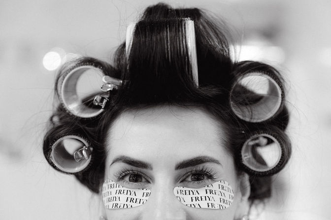 Close-up black-and-white photo of a woman wearing hair rollers and printed under-eye gel patches in a beauty salon.