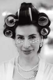 Black-and-white close-up of a woman in hair rollers and printed under-eye patches, smiling with pearl necklaces and earrings during a bridal getting-ready moment.
