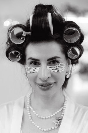 Black-and-white close-up of a woman in hair rollers and printed under-eye patches, smiling with pearl necklaces and earrings during a bridal getting-ready moment.