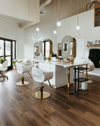 Bright modern hair salon interior with white-and-gold styling stations, oval mirrors, pendant globe lights, large windows, and warm wood flooring