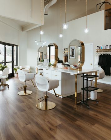Bright modern hair salon interior with white-and-gold styling stations, oval mirrors, pendant globe lights, large windows, and warm wood flooring