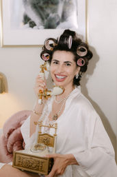 Smiling woman in a silk robe with pink hair rollers and pearl necklace holding a gold vintage telephone in a cozy boudoir