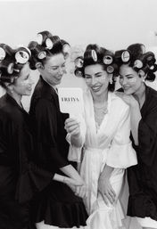 Bridal party getting-ready: four women in robes with hair rollers and under-eye patches laugh as the bride in a white robe holds a hand mirror.
