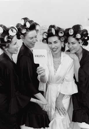 Bridal party getting-ready: four women in robes with hair rollers and under-eye patches laugh as the bride in a white robe holds a hand mirror.