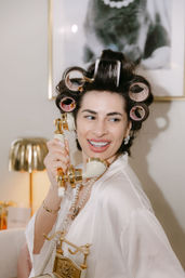 Smiling woman in hair rollers and a white silk robe holding a gold vintage telephone receiver, wearing pearl necklace and earrings in a glamorous salon-style getting-ready scene with a lamp and framed art in the background.
