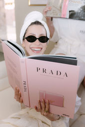 Smiling reader in a white spa robe with towel turban and tiny black sunglasses relaxes at home, holding a large pink Prada coffee-table book, showing gold rings and manicured nails.