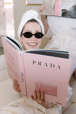 Smiling reader in a white spa robe with towel turban and tiny black sunglasses relaxes at home, holding a large pink Prada coffee-table book, showing gold rings and manicured nails.