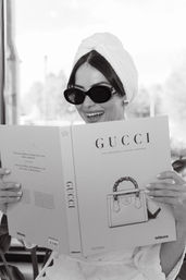 Black-and-white photo of a smiling woman in a towel turban and sunglasses reading a luxury handbag coffee-table book by a sunlit window.