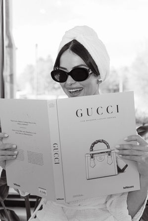 Black-and-white photo of a smiling woman in a towel turban and sunglasses reading a luxury handbag coffee-table book by a sunlit window.