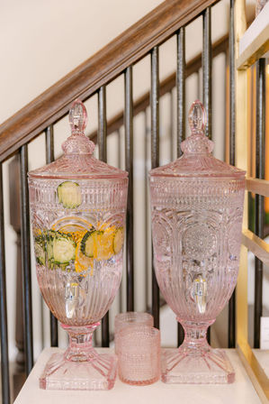 Pink ornate cut-glass beverage dispensers with spigots, one filled with lemon and cucumber slices, paired with matching pink tumblers on a white surface by a wooden staircase balustrade.