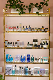 Gold metal shelving against a blush-pink wall showcasing organized skincare and haircare bottles, jars and boxed products on three illuminated tiers with potted trailing plants on top — chic boutique beauty display.