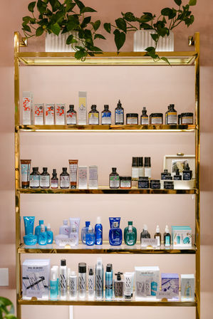 Gold metal shelving against a blush-pink wall showcasing organized skincare and haircare bottles, jars and boxed products on three illuminated tiers with potted trailing plants on top — chic boutique beauty display.