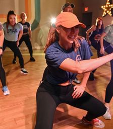 Energetic group dance fitness class in an indoor studio, smiling woman in a coral cap and athletic wear leading participants on a wooden floor
